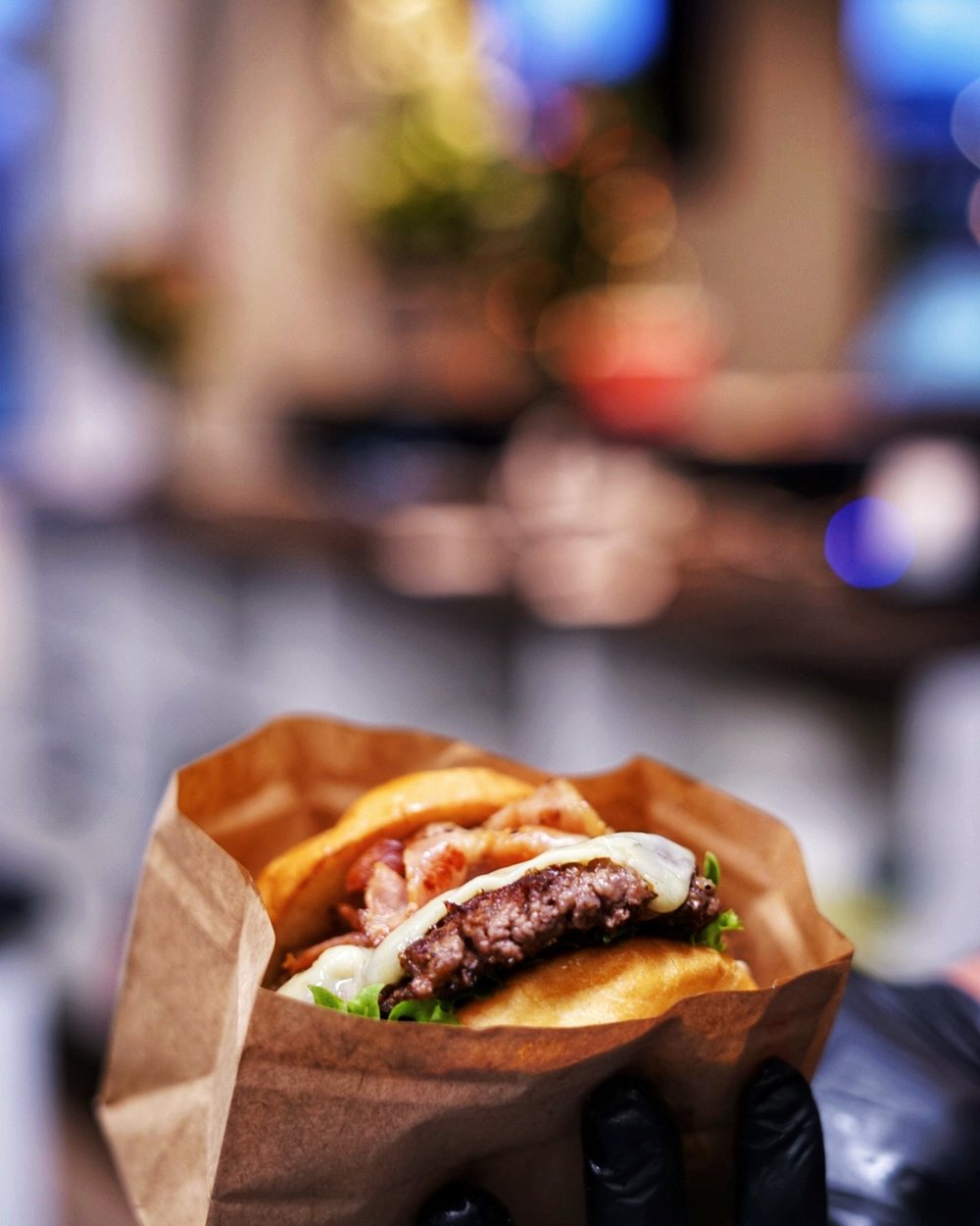 En smashburgare med bacon, smält ost och sallad i en papperspåse, hållen av en hand i svart handske